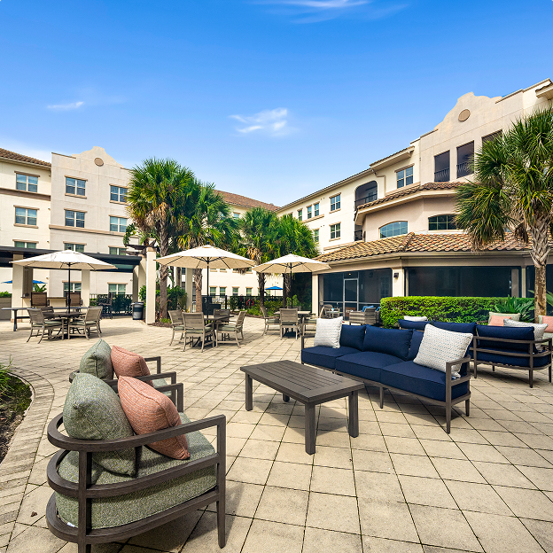 A spacious courtyard with outdoor seating, tables with umbrellas, palm trees, and a light-colored three-story building under a clear blue sky.