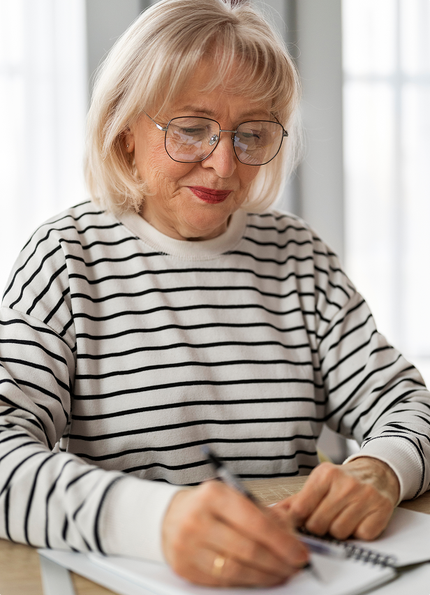 A senior woman with gray hair and glasses, wearing a black and white striped sweater, is sitting at a table and writing in a notebook with a pen. She looks focused and is smiling gently.