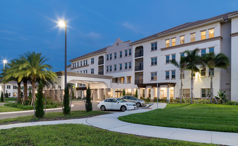 A modern four-story apartment building with lit windows at dusk, palm trees in front, and two parked cars near a covered entrance and a well-maintained lawn.