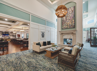 Bright, modern lounge area with a sofa, two wicker chairs, a coffee table, a large fireplace, and contemporary artwork. A dining area with tables and chairs is visible in the background.
