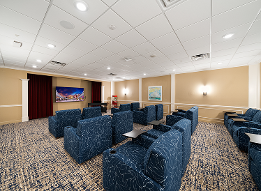 A small theater room with patterned blue armchairs, tray tables, a large screen at the front, beige walls, framed artwork, and soft lighting from wall sconces.
