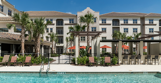 A resort-style apartment complex features a swimming pool, lounge chairs, shaded pergolas, palm trees, and outdoor seating with orange umbrellas under a clear blue sky.
