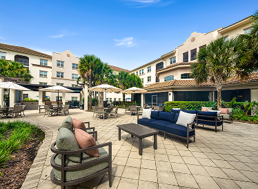 Outdoor courtyard with cushioned seating, tables, umbrellas, and palm trees, surrounded by a beige multi-story building under a clear blue sky.