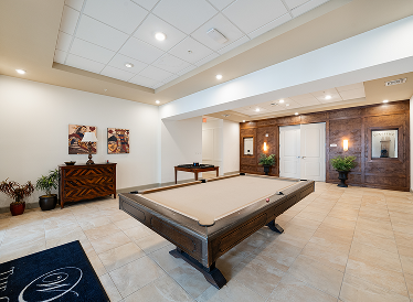 A spacious, well-lit game room with a beige pool table in the center, wood-paneled accent wall, potted plants, artwork, and a small table with games. The floor is tiled, and the ceiling has recessed lighting.