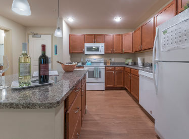 Modern kitchen with wooden cabinets, granite countertops, white appliances, and a center island. Two bottles of wine and a wine glass are on the counter, with a calendar and notes on the refrigerator.