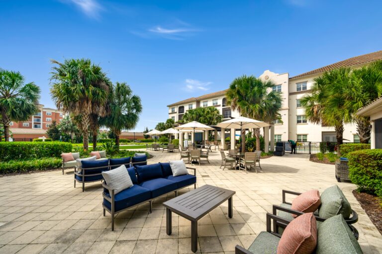 Spacious outdoor patio with cushioned chairs, sofas, and tables under umbrellas, surrounded by palm trees and landscaping, adjacent to a white multi-story building under a clear blue sky.