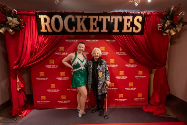 A smiling woman in a green costume poses next to a senior with a cane in front of a red ROCKETTES sign and a Radio City Music Hall backdrop decorated with red curtains and holiday ornaments.