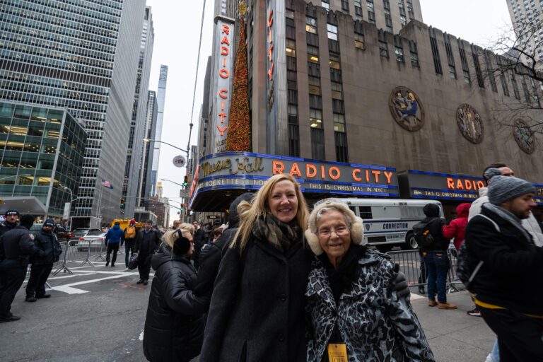 Two women smiling and posing for a photo outside Radio City Music Hall in New York City, with people walking and city buildings in the background.