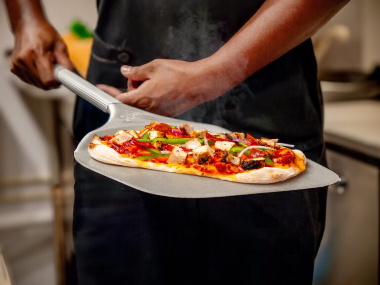 A person holds a pizza peel with a freshly baked pizza topped with cheese, chicken, and vegetables in a kitchen setting. Steam rises from the hot pizza.