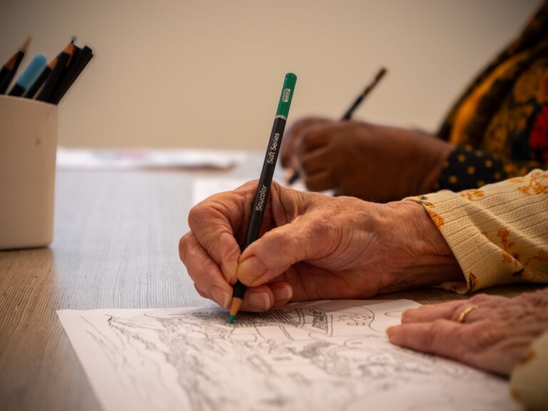 Close-up of two people coloring in a detailed drawing with colored pencils at a table. One person wears a patterned shirt and a ring; the other has darker skin and holds a pencil, partially visible in the background.