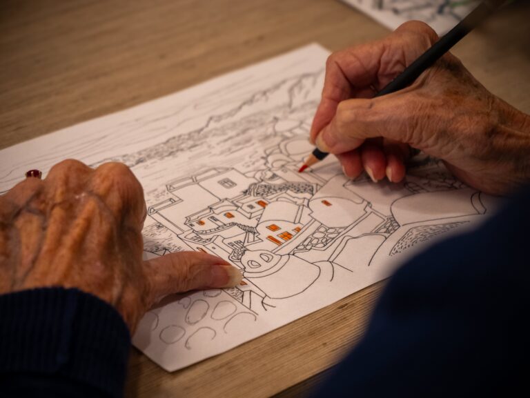 Close-up of elderly hands coloring a detailed line drawing of houses and landscape with an orange pencil on a wooden table. The person is wearing a dark blue sleeve.