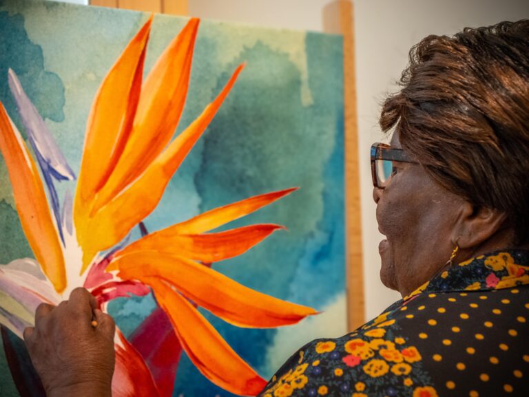 A senior woman with glasses and a polka-dot shirt paints a vibrant orange and yellow flower on a canvas with a blue-green background. She is focused on her artwork, which features bold colors and expressive brushstrokes.