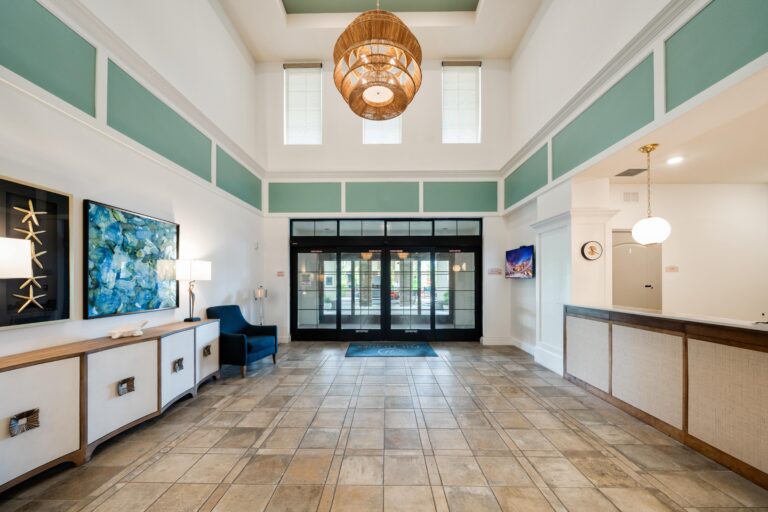 A bright, spacious hotel lobby with tiled floors, high ceilings, modern decor, a chandelier, seating area, front desk, and large glass entrance doors letting in natural light.