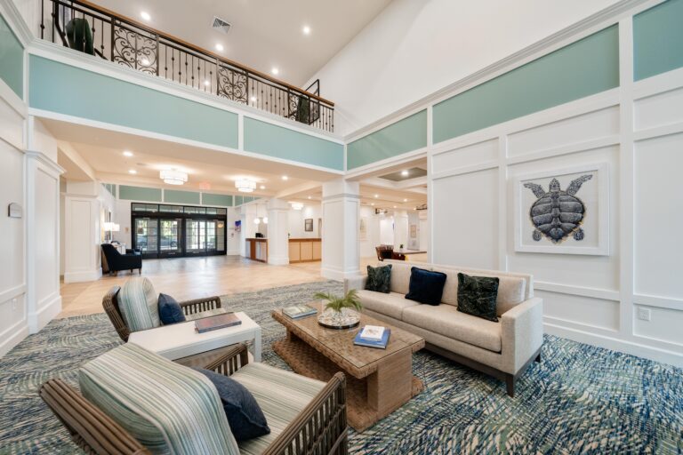 Bright, modern lobby with high ceilings, a balcony, and elegant decor. Features a sofa, armchairs, coffee table, wall art of a turtle, blue-green accents, and large windows letting in natural light.