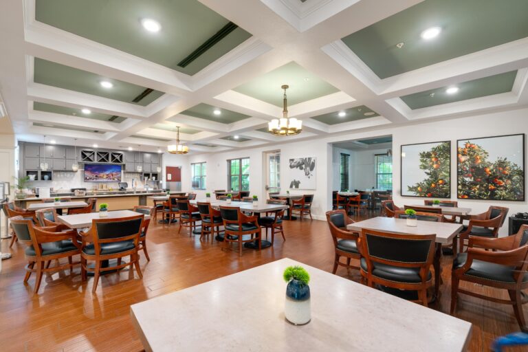 A spacious, well-lit dining area with wooden tables and cushioned chairs, decorated with green plants and large wall art, featuring a modern kitchen in the background.