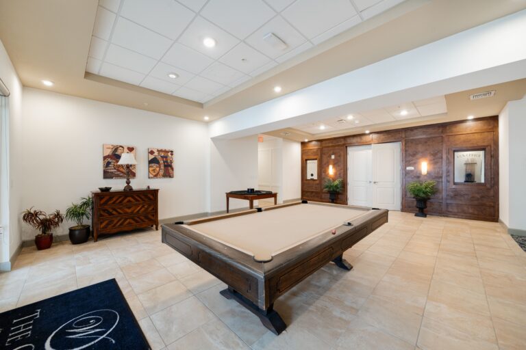 A spacious game room with a pool table in the center, light tiled floors, wood-paneled walls, potted plants, artwork, and a small side table near the entrance. Ceiling lights provide bright, even illumination.