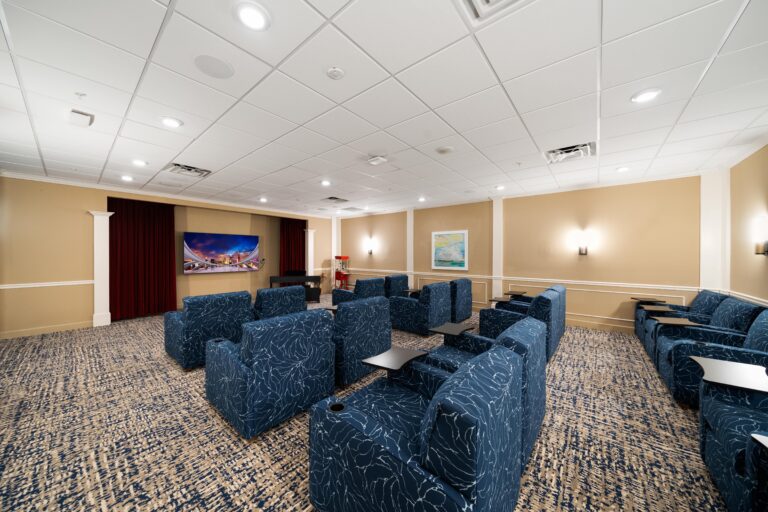 A small theater room with rows of blue patterned armchairs facing a wall-mounted TV, beige walls, framed artwork, and carpeted flooring. Ceiling lights and side wall sconces provide illumination.