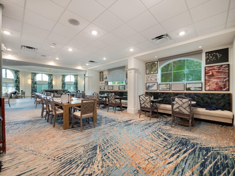 Spacious, modern dining area with wooden tables and chairs, patterned carpet, large windows letting in natural light, and framed artwork on the walls. The space has a clean, bright, and inviting atmosphere.