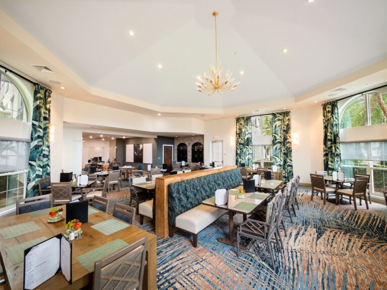 Bright, modern restaurant interior with large windows, patterned curtains, stylish lighting, and a mix of booth and table seating. Tables are set with menus, napkins, and small flower arrangements.