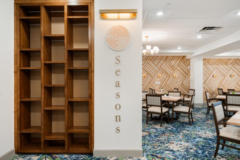 A modern dining area with patterned wallpaper, wooden chairs around tables, and a large empty wooden shelving unit. The wall features a circular tree design with the word Seasons displayed vertically.