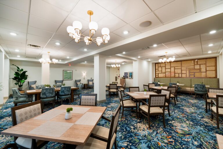 A spacious, well-lit dining area with multiple tables and cushioned chairs, chandeliers hanging from the ceiling, patterned blue carpet, and modern decor with plants and wall art.