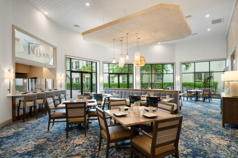 A modern, spacious dining area with wooden tables and chairs, large windows, blue patterned carpet, pendant lights, and a sign reading La Boon above a counter. Greenery is visible outside.