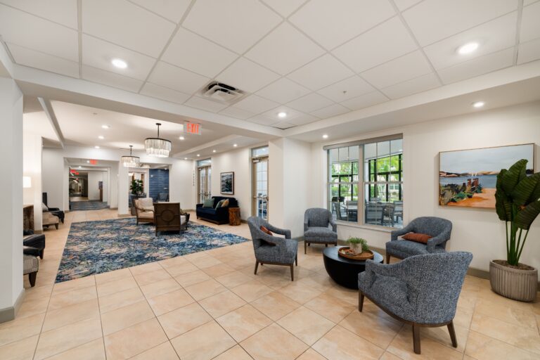 A spacious, modern lounge area with four gray armchairs around a small table, large windows, tile floors, potted plants, artwork on the walls, and a hallway with additional seating in the background.