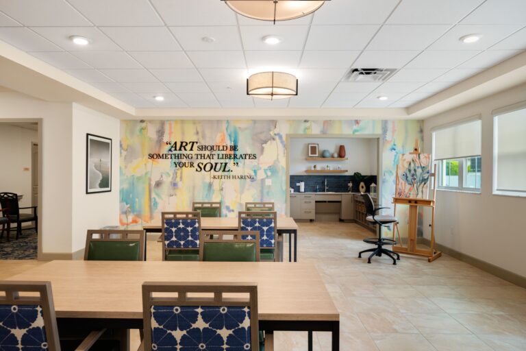 A bright, modern art room with tables and chairs, colorful wall art featuring a quote by Keith Haring, shelves with art supplies, windows letting in natural light, and an easel near the back wall.