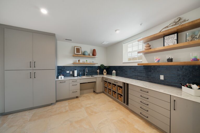 Modern, spacious laundry or craft room with gray cabinets, black tile backsplash, open wooden shelves with decor, baskets, and supplies, tan tile floor, and a large countertop under a window.