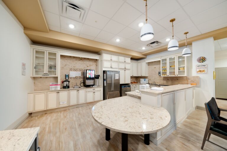 Spacious, modern kitchen with cream cabinets, stainless steel fridge, coffee and soda machines, marble countertops, and pendant lights hanging from a white tiled ceiling. A round table and black chairs sit in the center.