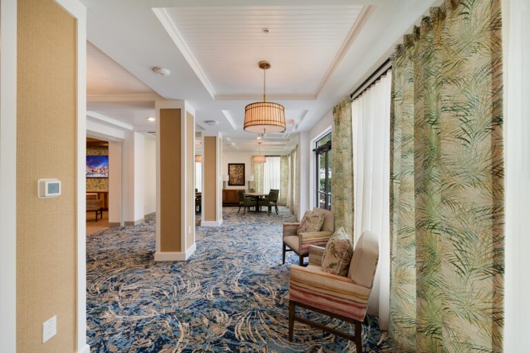 A modern, well-lit lounge area with patterned blue carpet, two upholstered chairs near tall windows with leafy curtains, and ceiling lights. In the background, tables and wall art are visible.