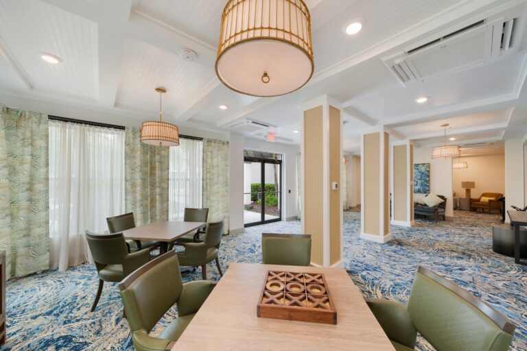 A bright, modern lounge with green chairs, wooden tables, patterned blue carpet, large windows with sheer curtains, and circular pendant lights. A tic-tac-toe game is set on the nearest table.