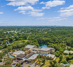 Aerial view of a large building complex surrounded by trees, small lakes, and greenery, with a sprawling suburban neighborhood and blue sky in the background.