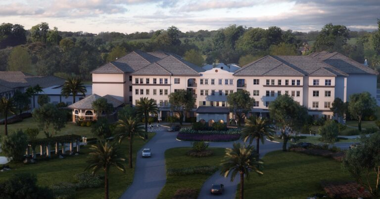 A large, elegant white building with a peaked roof sits amid landscaped gardens, palm trees, and winding driveways, surrounded by lush greenery and distant trees under a partly cloudy sky.