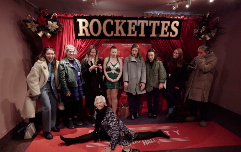 A group poses in front of a red curtain with a large ROCKETTES sign. A woman in the front does a split while others smile and stand behind her, including someone dressed as a Rockette. Festive decorations frame the scene.