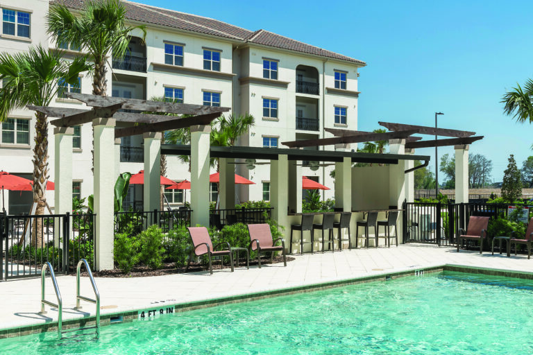 Outdoor swimming pool with lounge chairs and a shaded seating area in front of a modern, four-story apartment building. Palm trees and red umbrellas provide a resort-like atmosphere under a clear blue sky.
