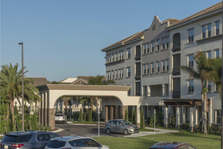 A modern multi-story building with arched entryway and balconies, surrounded by palm trees and parked cars, under a clear blue sky.