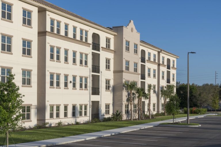 A modern, four-story apartment building with beige and white exterior, numerous windows, and palm trees in front. The parking lot in the foreground is empty under a clear blue sky.
