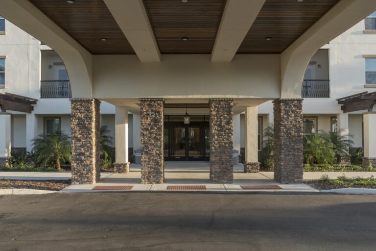 Covered entrance of a modern building with stone pillars, glass doors, and landscaped plants on either side, viewed from the driveway.