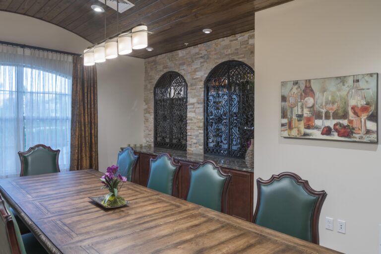 A dining room with a long wooden table, green upholstered chairs, a ceiling light fixture, two decorative wall niches with metal grilles, a painting of wine bottles and glasses, and a vase with purple flowers on the table.