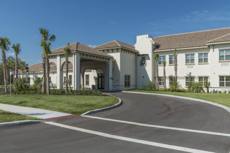 A modern, two-story building with a tile roof and arched entrance, surrounded by palm trees and landscaping, sits under a clear blue sky with a curved driveway leading to the front.