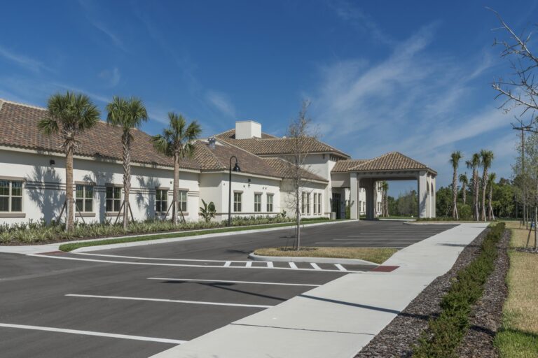 A modern, single-story building with a tiled roof and covered entrance is surrounded by palm trees and landscaping. The empty parking lot and clear blue sky suggest a sunny day.
