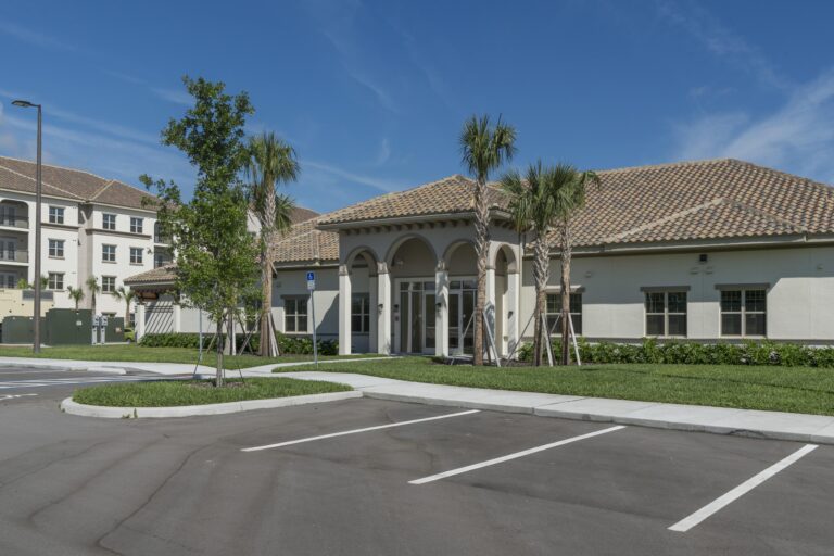 A building with a tiled roof and arched entrance is surrounded by palm trees and landscaping, with an empty parking lot in front, under a clear blue sky.