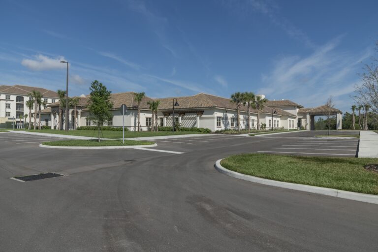 A modern, single-story building with a tiled roof stands next to an empty parking lot under a blue sky with light clouds and surrounding green lawns and palm trees.