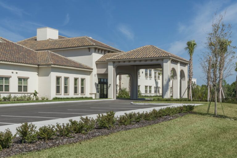 A modern beige building with a tiled roof, large arches at the entrance, several windows, and a mostly empty parking lot, surrounded by green grass, shrubs, and young trees under a clear blue sky.