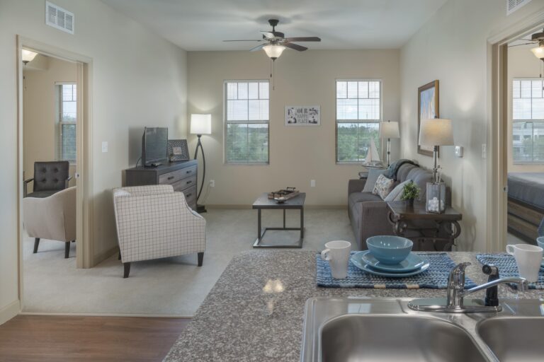 Modern apartment living room with neutral tones, seen from the kitchen. Features a sofa, armchairs, TV, decor, and natural light from multiple windows. A dining set and sink are visible in the foreground.