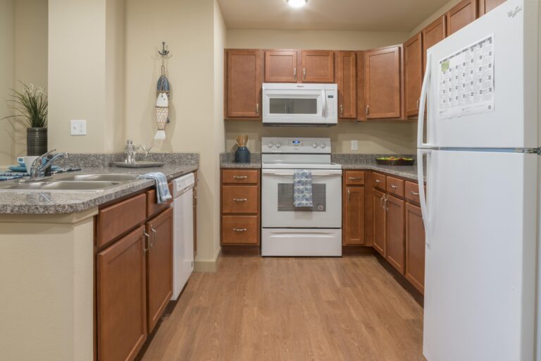 A modern kitchen with wooden cabinets, white appliances including a refrigerator, stove, microwave, and dishwasher, and a double sink. Counters have minimal decor and a dish towel hangs by the sink.