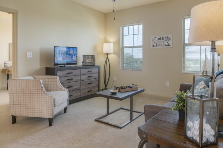 A cozy living room with beige walls and carpet, a cushioned armchair, a dresser with a TV, a coffee table, a sofa, decorative plants, and windows letting in natural light.