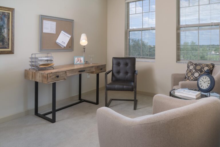 A tidy home office with a wooden desk, metal chair, and beige armchair. The desk holds a lamp, file organizer, and photo. A round glass side table displays a clock. Windows display a scenic outdoor view.