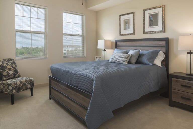 A neatly made bed with blue bedding and pillows sits between two lamps on nightstands in a bright bedroom with beige walls, two windows, framed art, and an upholstered chair in the corner.